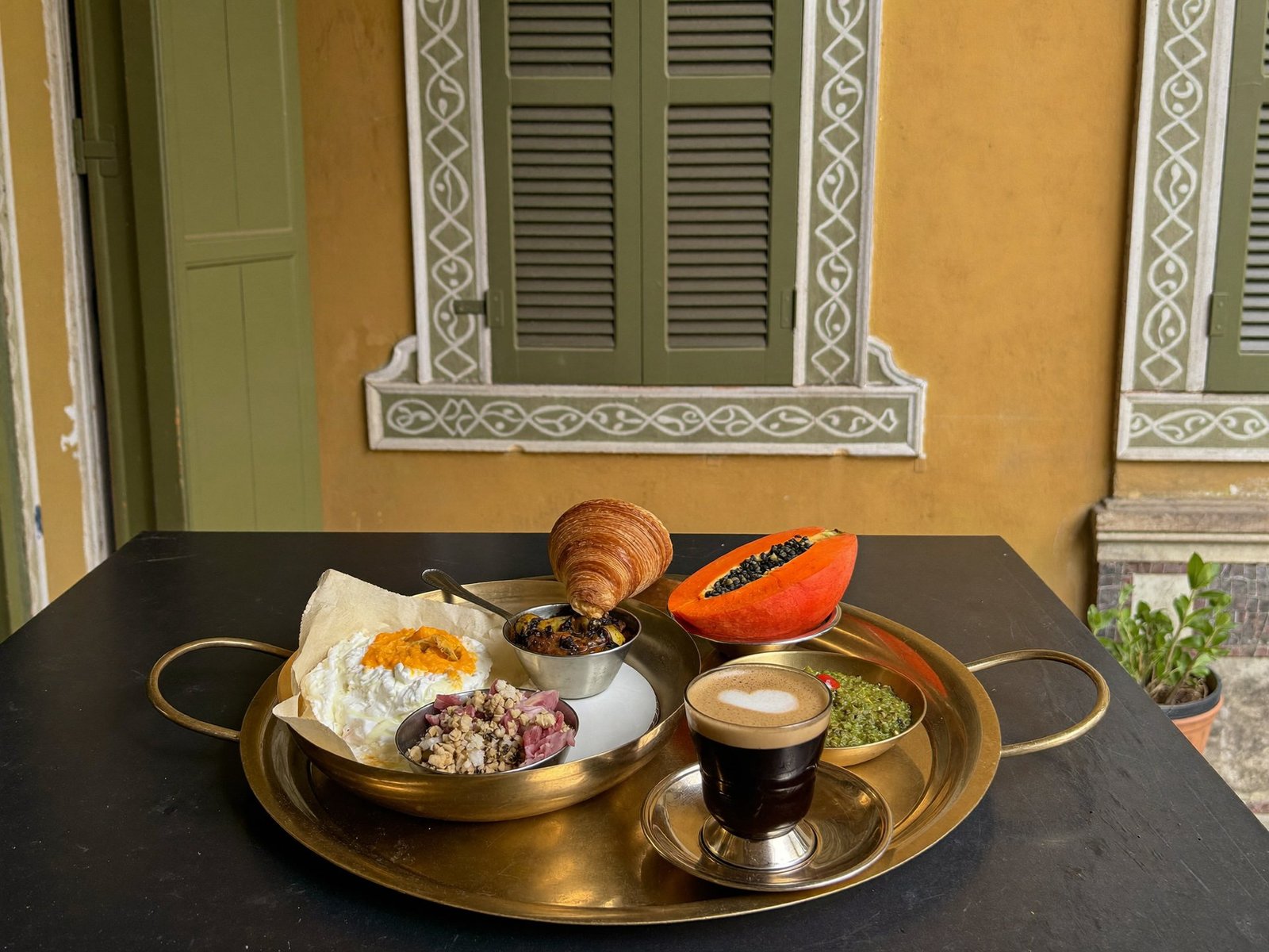 A brass thali breakfast served on a dark table beside green louvred shutters